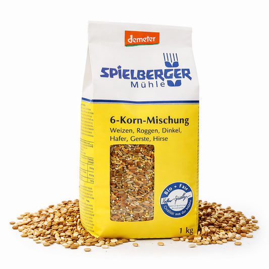 Packung Spielberger Demeter 6-Korn-Mischung 1000g stehend, umgeben von verstreuten Körnern, Vorderseite mit Sichtfenster und gelbem Etikett sichtbar.