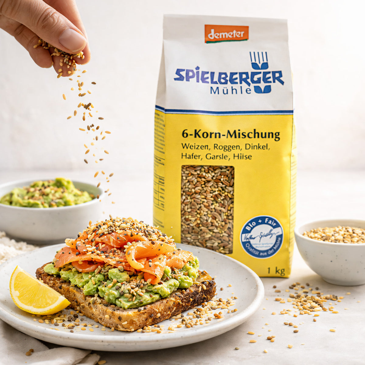 Hand streut Spielberger Korn-Mischung über belegtes Brot mit Avocado und Lachs, Packung 6-Korn-Mischung und Schale mit Körnern im Hintergrund, Frühstücksszene.