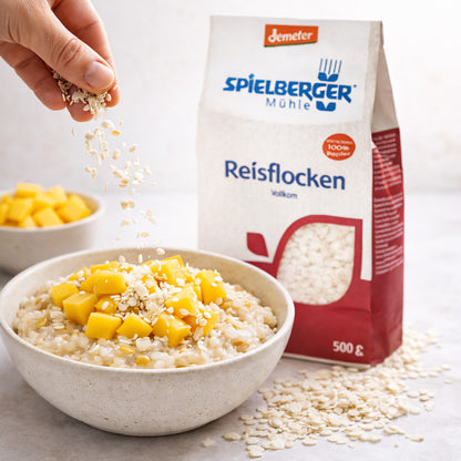 Hand streut Spielberger Reisflocken über eine Frühstücksschale mit Reisflocken und Mangostücken, Packung im Hintergrund sichtbar.