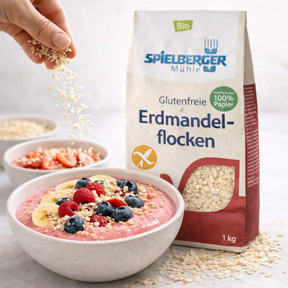 Hand streut erdmandelflocken von spielberger über eine Smoothie-Bowl mit Beeren und Bananen, Verpackung im Hintergrund.