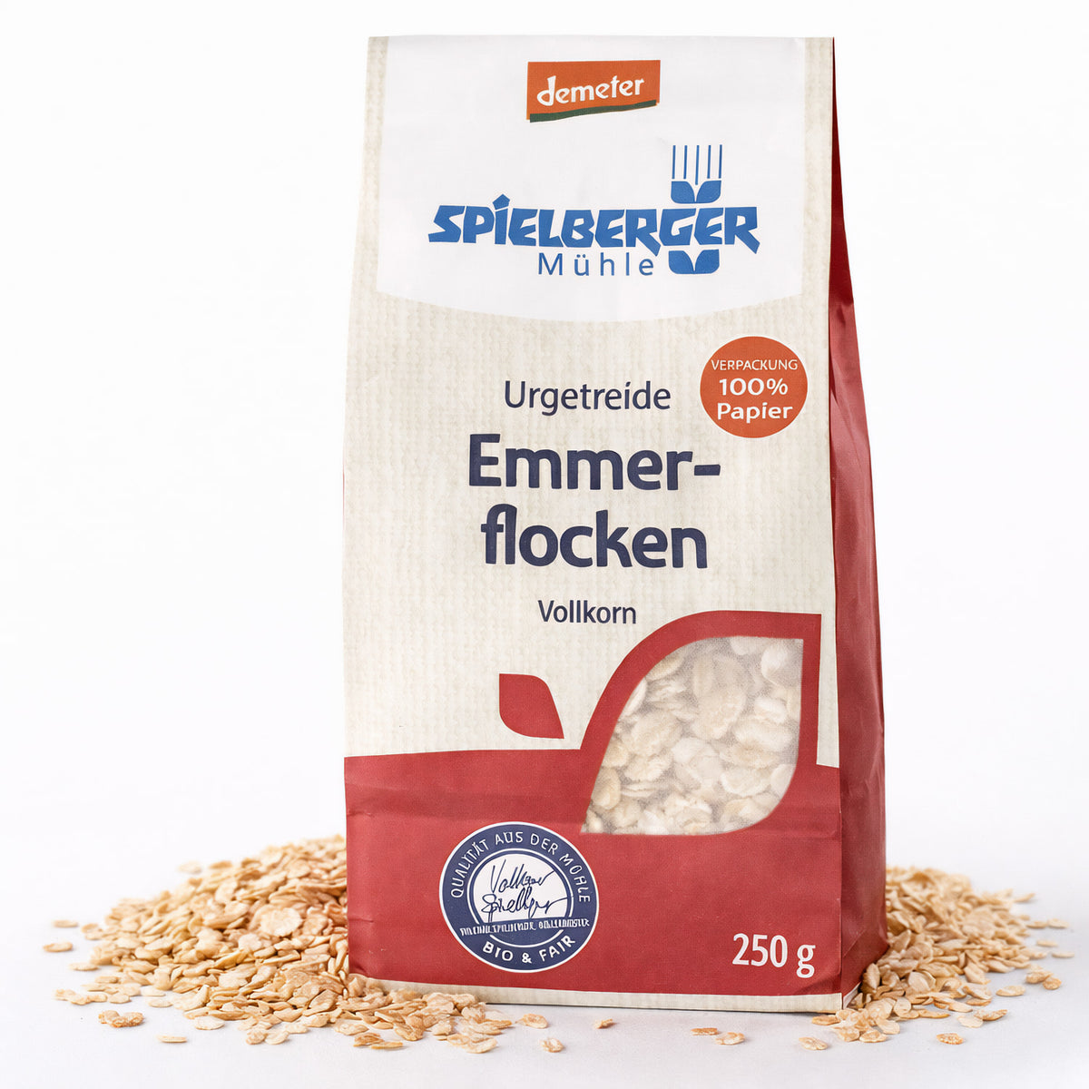 spielberger Emmerflocken demeter 250g in Papierverpackung, stehend auf weißem Untergrund mit verstreuten Emmerflocken davor