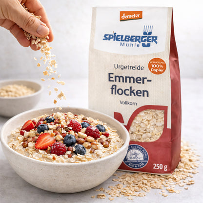 schale mit Müsli aus emmerflocken von spielberger, garniert mit Beeren und Nüssen, daneben die demeter 250g Packung, Hand streut Flocken darüber