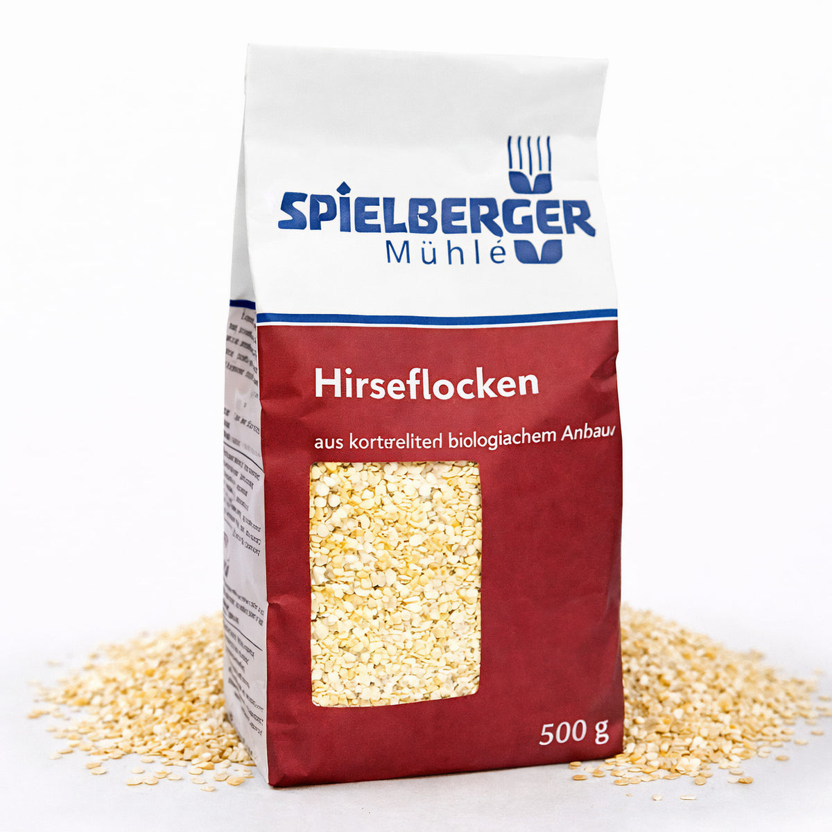 spielberger hirseflocken 500g in roter Verpackung mit Sichtfenster, umgeben von verstreuten Hirseflocken auf weißem Untergrund