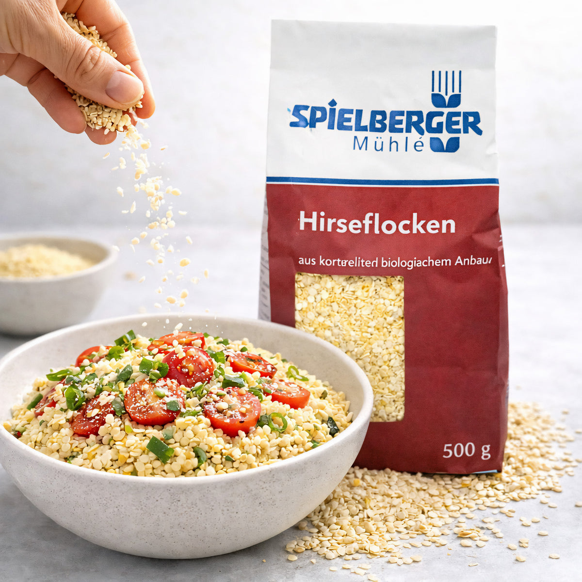 hirseflocken von spielberger werden über eine Schüssel mit Tomaten und Frühlingszwiebeln gestreut, Verpackung und weitere Schale im Hintergrund