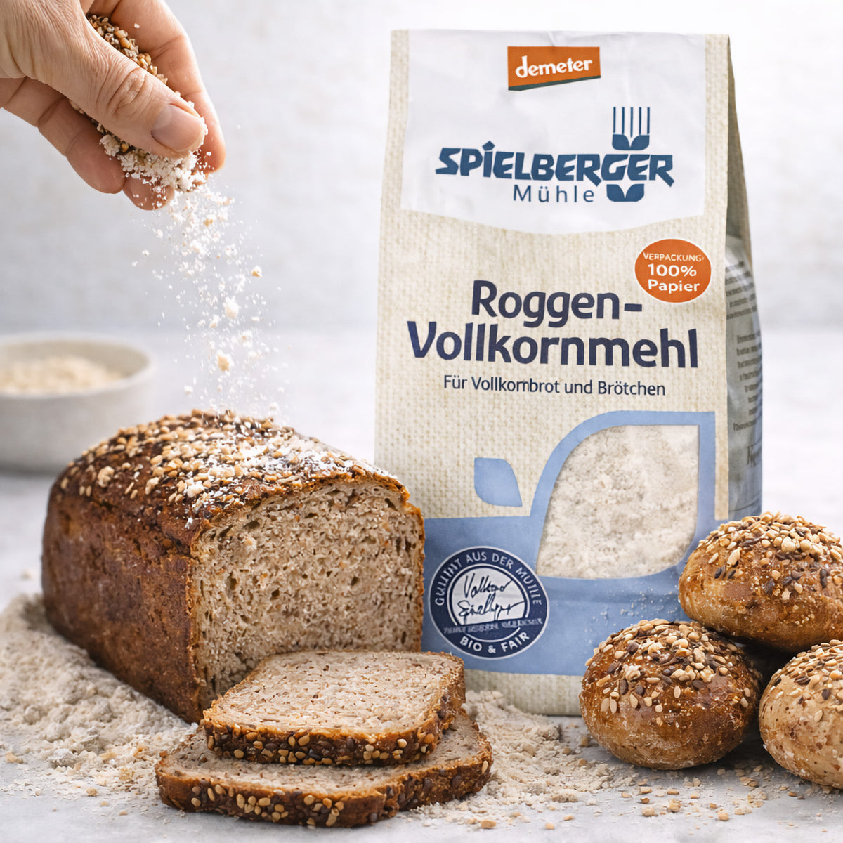 Spielberger Roggen-Vollkornmehl demeter neben Roggenbrot und Brötchen, eine Hand streut Körner auf das Brot, im Hintergrund eine Schale.