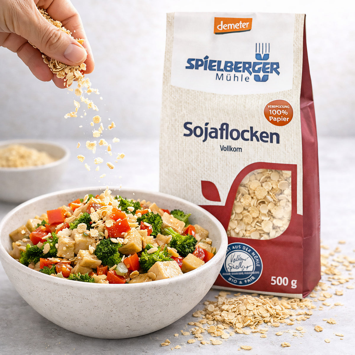 Hand streut spielberger sojaflocken über eine Schüssel mit Gemüse, im Hintergrund die demeter 500g Packung, geeignet für frühstück oder snacks.