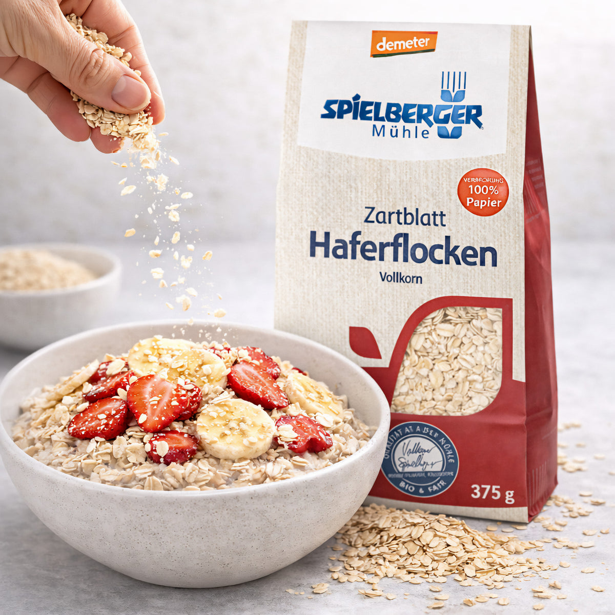 Schale mit Haferflocken Zartblatt von Spielberger, belegt mit Erdbeeren und Bananen, daneben die geöffnete Packung, Haferflocken werden von Hand darüber gestreut.