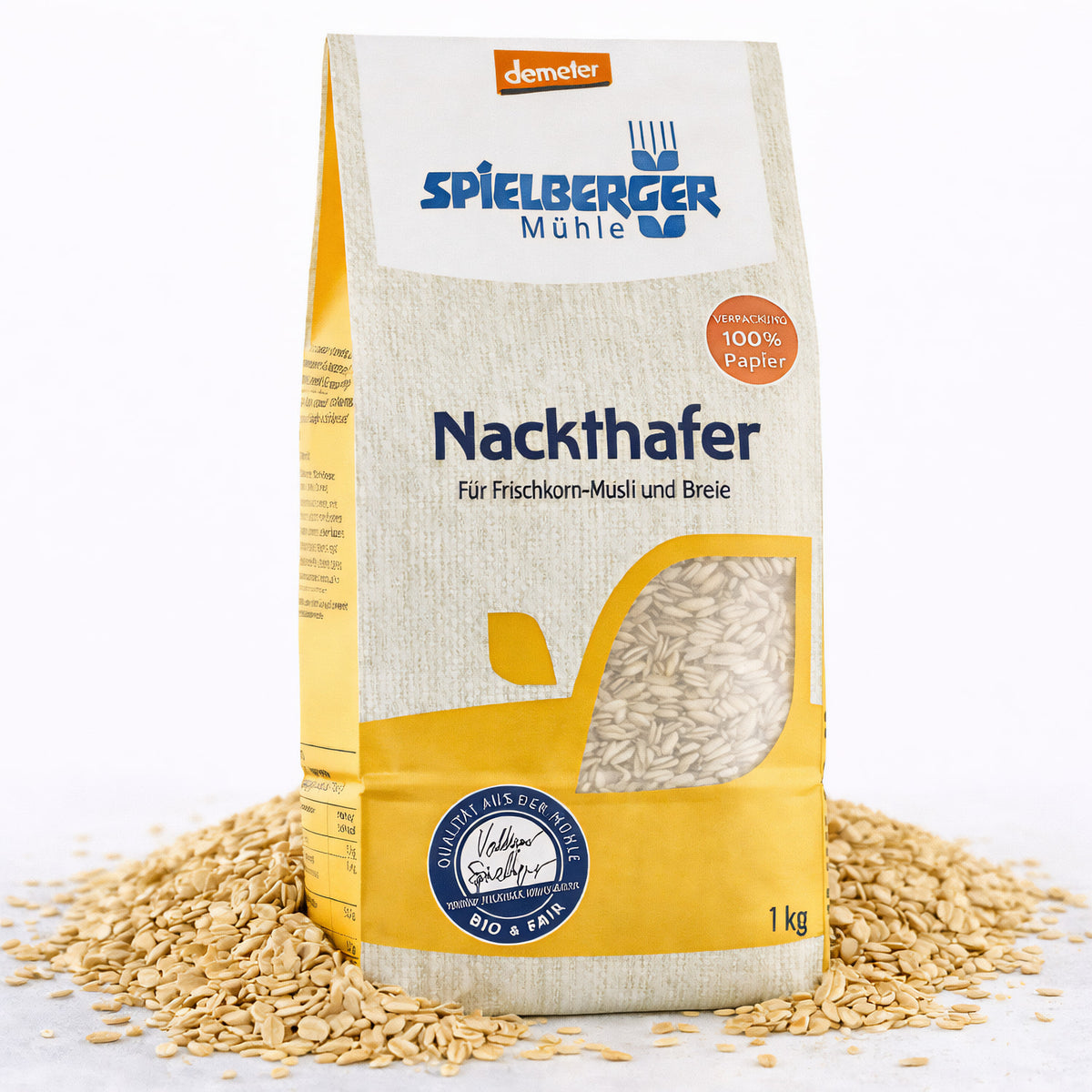 Packung Spielberger Nackthafer, demeter, 1 kg, stehend auf einer Schicht loser Haferkörner, Vorderseite mit Sichtfenster und Produktinformationen sichtbar.
