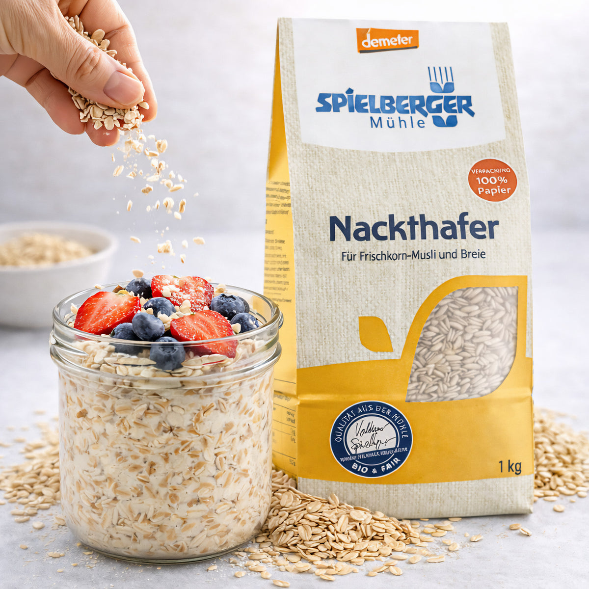 Hand streut Spielberger Nackthafer über ein Glas mit Haferflocken, Erdbeeren und Heidelbeeren, Packung Nackthafer demeter im Hintergrund, Haferkörner auf dem Tisch verteilt.