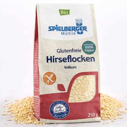 Spielberger glutenfreie Hirseflocken 250g in Papierverpackung, stehend auf verstreuten Flocken, Vorderseite mit Produktinfos sichtbar.