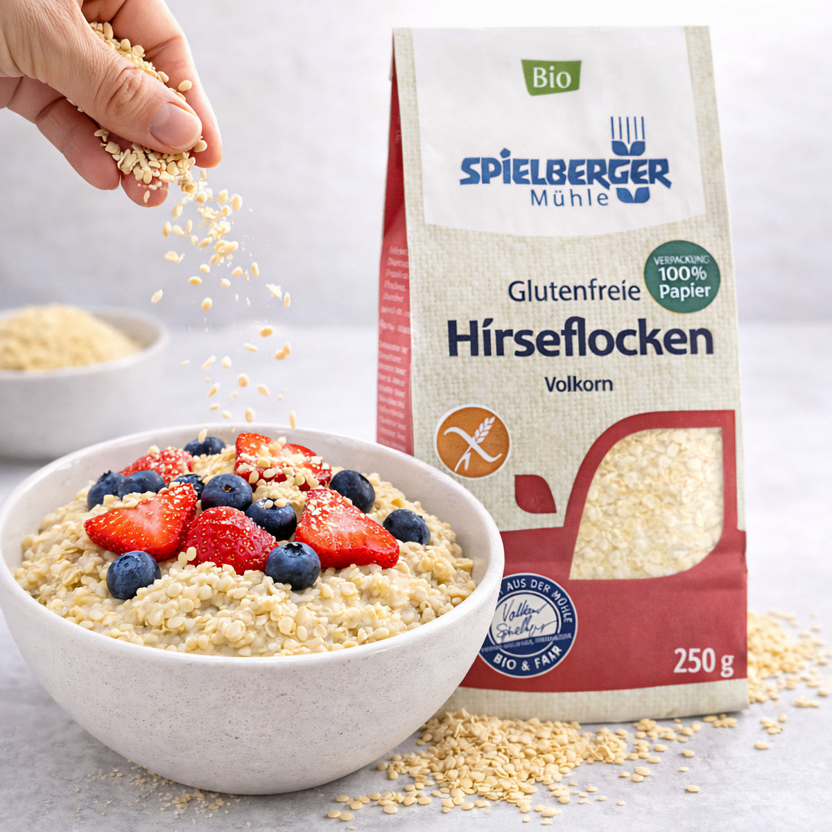 Schale mit Hirseflocken als Frühstück, garniert mit Erdbeeren und Heidelbeeren, daneben Spielberger glutenfreie Hirseflocken 250g, Hand streut Flocken als Topping darüber.