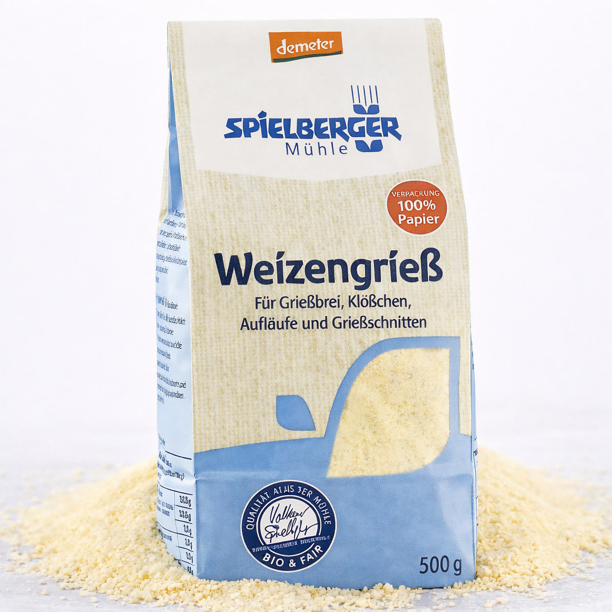 Eine 500g-Packung Spielberger Weizengrieß steht auf einer Unterlage aus losem Grieß, Vorderseite mit Produktinformationen sichtbar.