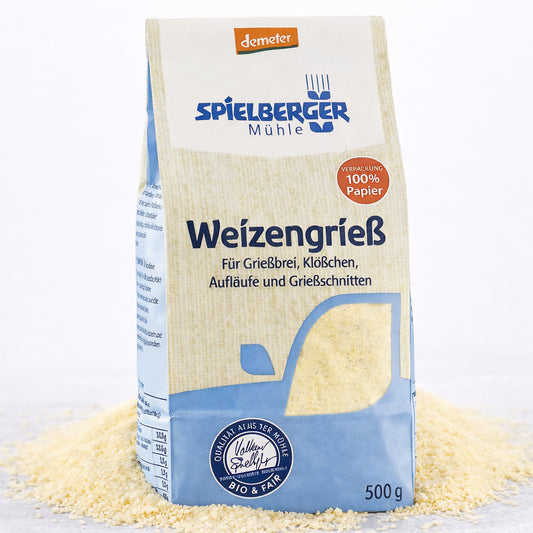 Eine 500g-Packung Spielberger Weizengrieß steht auf einer Unterlage aus losem Grieß, Vorderseite mit Produktinformationen sichtbar.