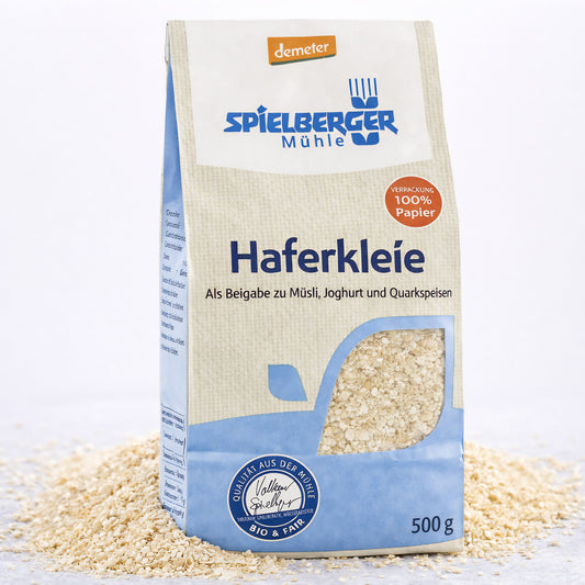 Spielberger Haferkleie demeter 500g Beutel stehend, umgeben von verstreuter Haferkleie auf hellem Untergrund.