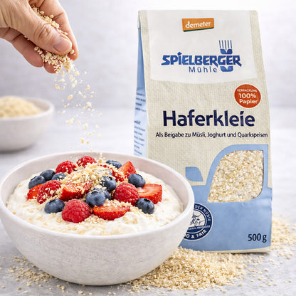 Hand streut Spielberger Haferkleie über eine Frühstücksschale mit Joghurt und frischen Beeren, daneben der 500g Beutel.