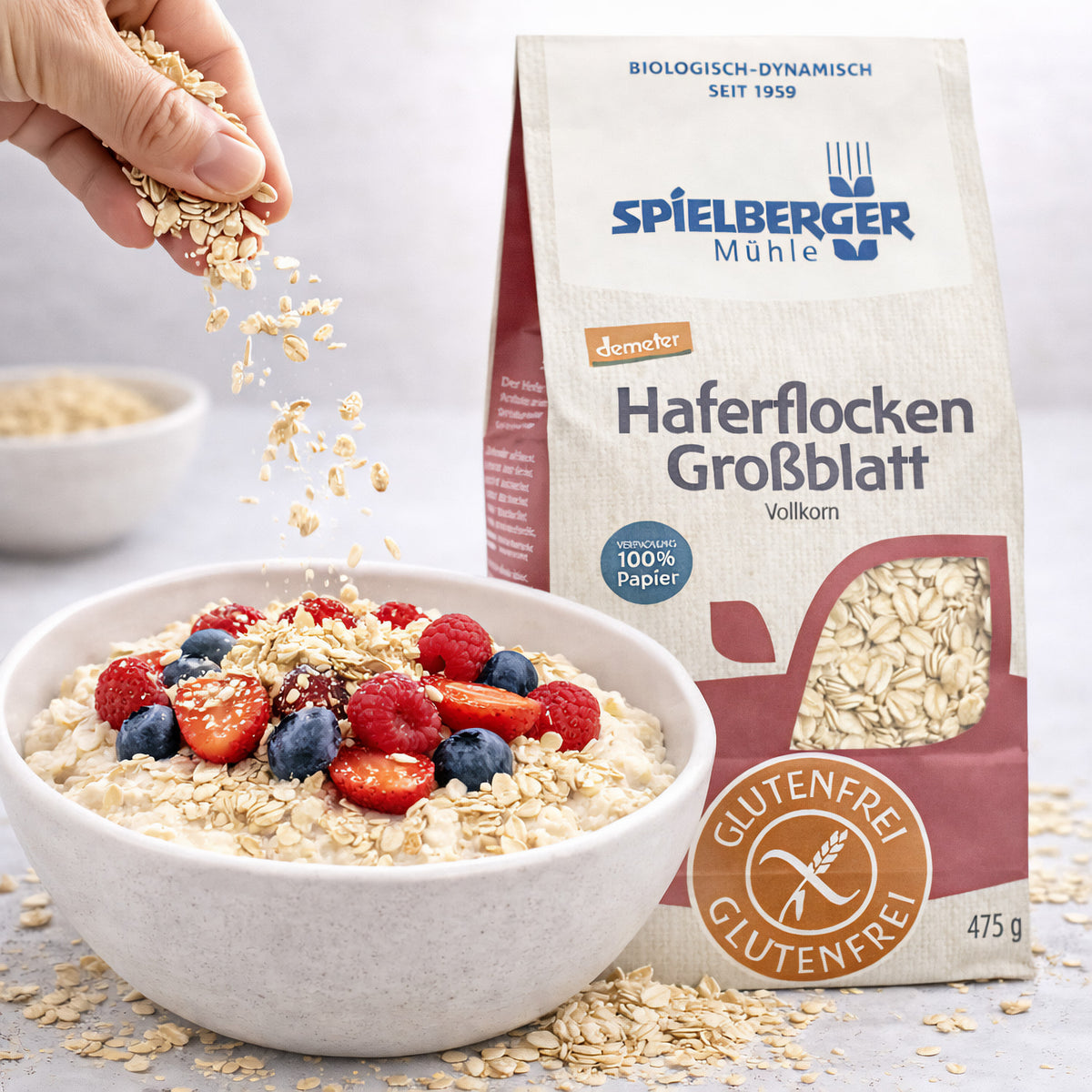 Hand streut glutenfreie Haferflocken Großblatt von Spielberger über eine Frühstücksschale mit Beeren, daneben die geöffnete Produktverpackung.