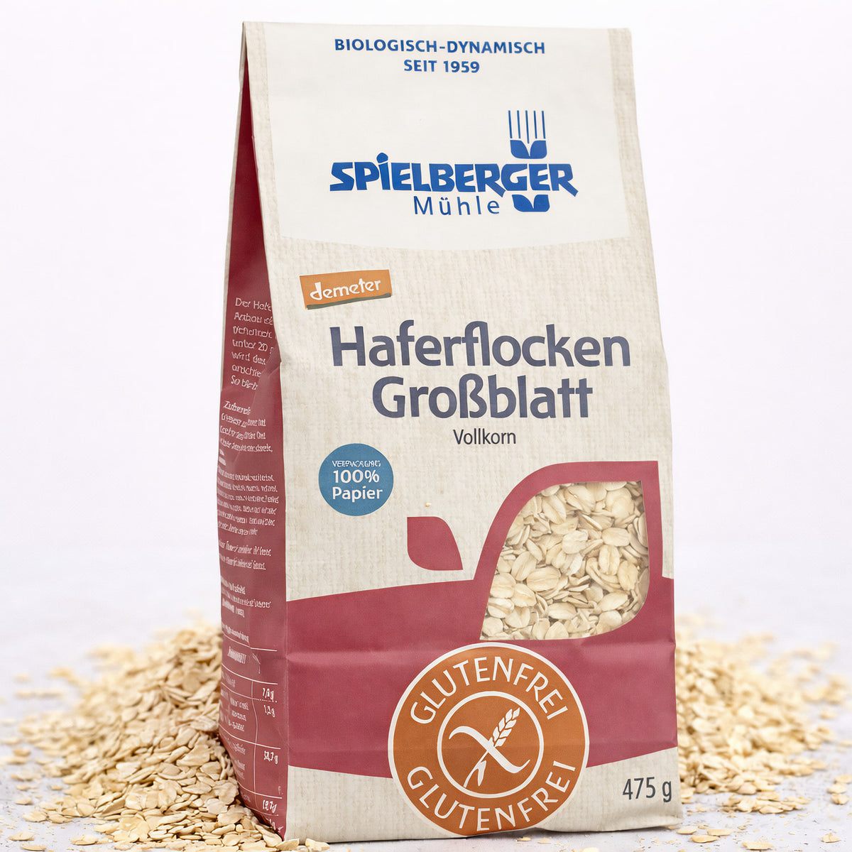 Spielberger glutenfreie Haferflocken Großblatt demeter 475g in Papierverpackung, stehend auf verstreuten Haferflocken, Vorderseite mit Sichtfenster und Produktinformationen.