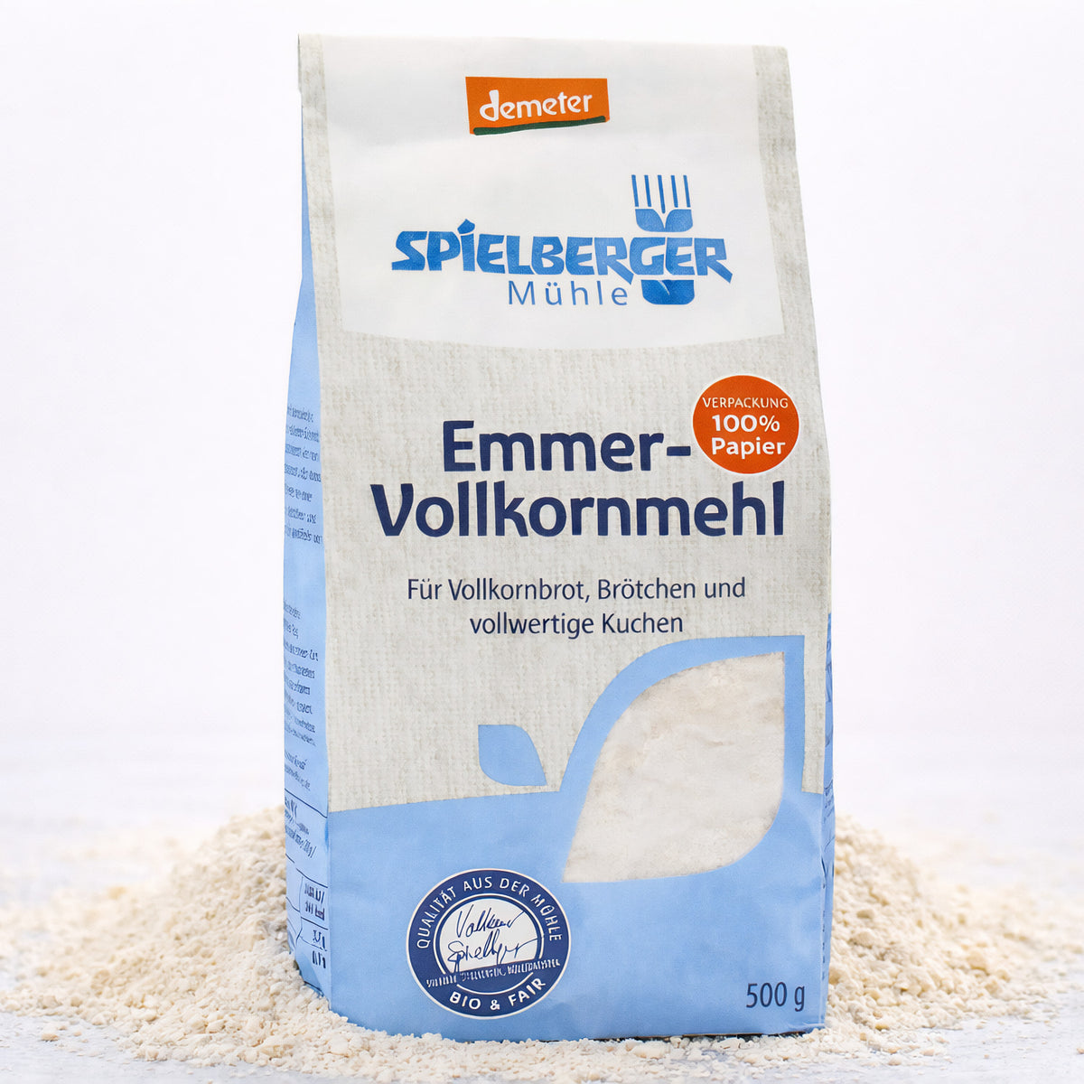 Eine stehende 500g-Packung spielberger Emmer-Vollkornmehl, umgeben von verstreutem Mehl auf hellem Untergrund.