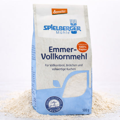 Eine stehende 500g-Packung spielberger Emmer-Vollkornmehl, umgeben von verstreutem Mehl auf hellem Untergrund.