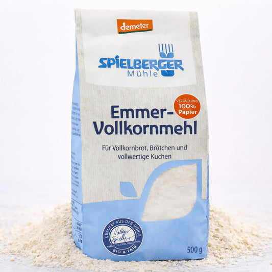 Eine stehende 500g-Packung spielberger Emmer-Vollkornmehl, umgeben von verstreutem Mehl auf hellem Untergrund.