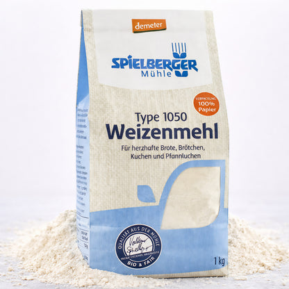 Eine stehende 1-kg-Packung spielberger Weizenmehl Type 1050, umgeben von verstreutem Mehl auf hellem Untergrund.