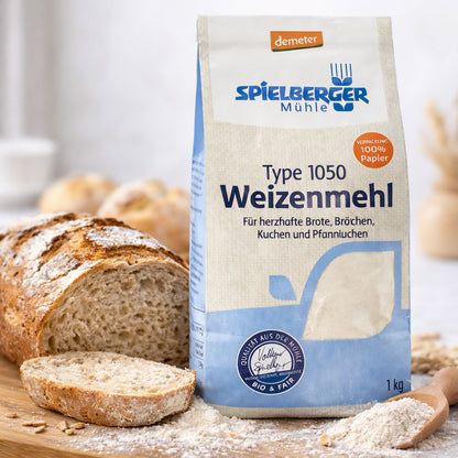 Eine Packung spielberger Weizenmehl 1050 steht auf einem Holzbrett neben angeschnittenem Brot und etwas verstreutem Mehl.