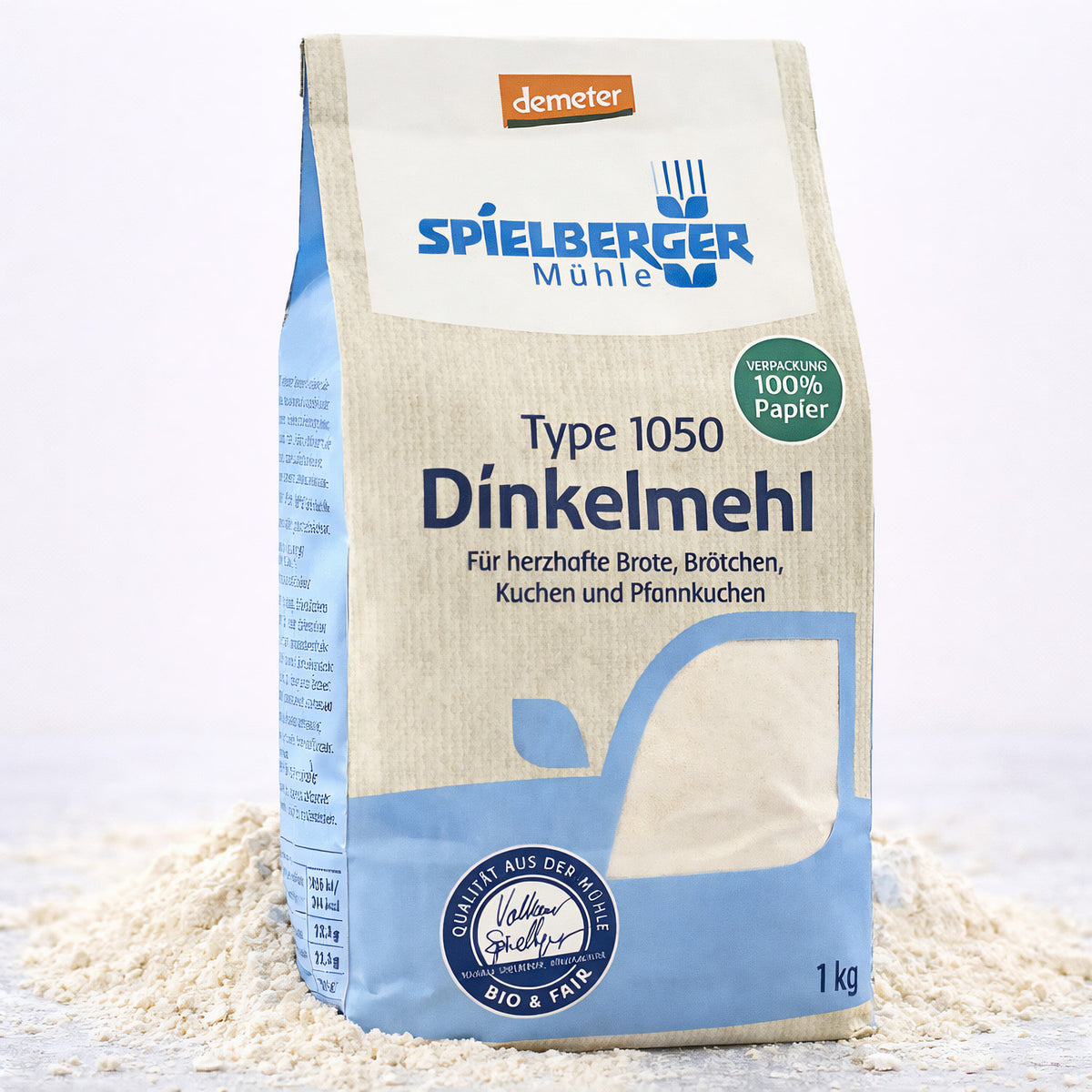 Eine stehende Packung Spielberger Dinkelmehl Type 1050, umgeben von verstreutem Mehl auf hellem Untergrund.