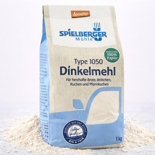 Eine stehende Packung Spielberger Dinkelmehl Type 1050, umgeben von verstreutem Mehl auf hellem Untergrund.