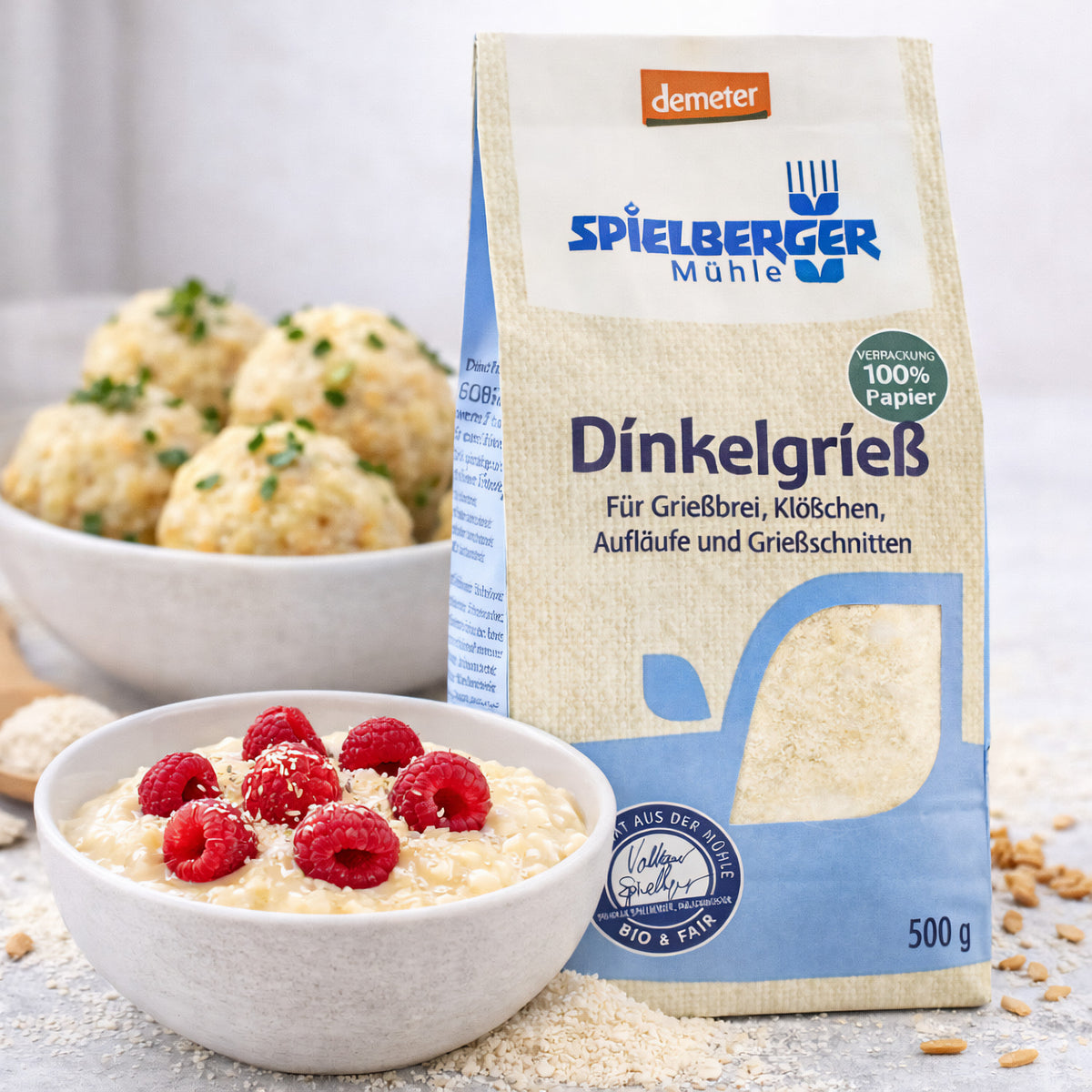 Spielberger Dinkelgrieß 500g neben einer Schale Grießbrei mit Himbeeren und einer weiteren Schale mit Klößchen, auf einem Tisch arrangiert.