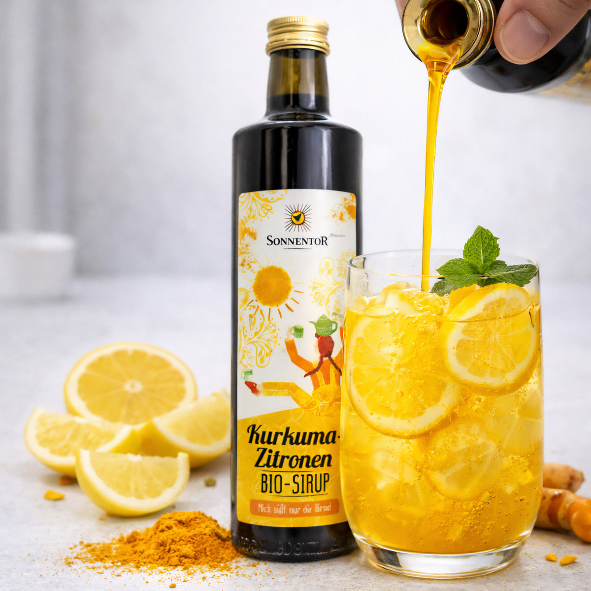 Kurkuma-Zitronen Sirup von Sonnentor wird in ein Glas mit Zitronenscheiben, Eiswürfeln und Minze gegossen, daneben frische Zitronen und Kurkuma.