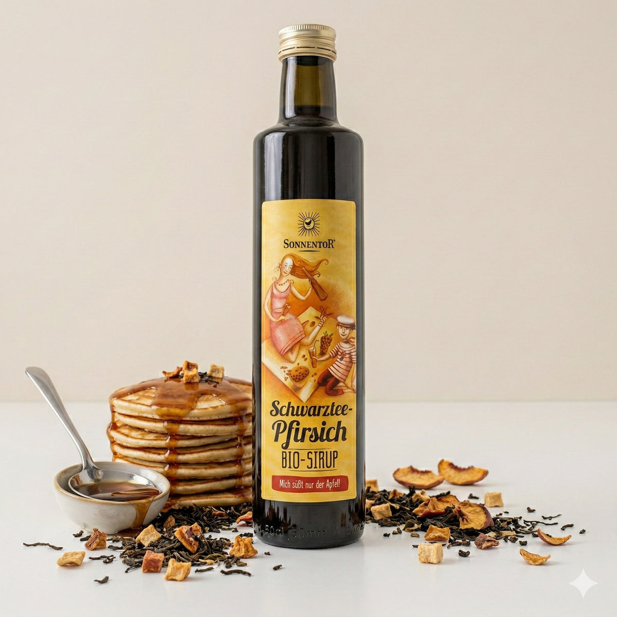 Sonnentor Schwarztee-Pfirsich Sirup 500ml vor einem Stapel Pancakes mit Sirup und Teeblättern sowie Pfirsichstücken, daneben ein Löffel mit Sirup.