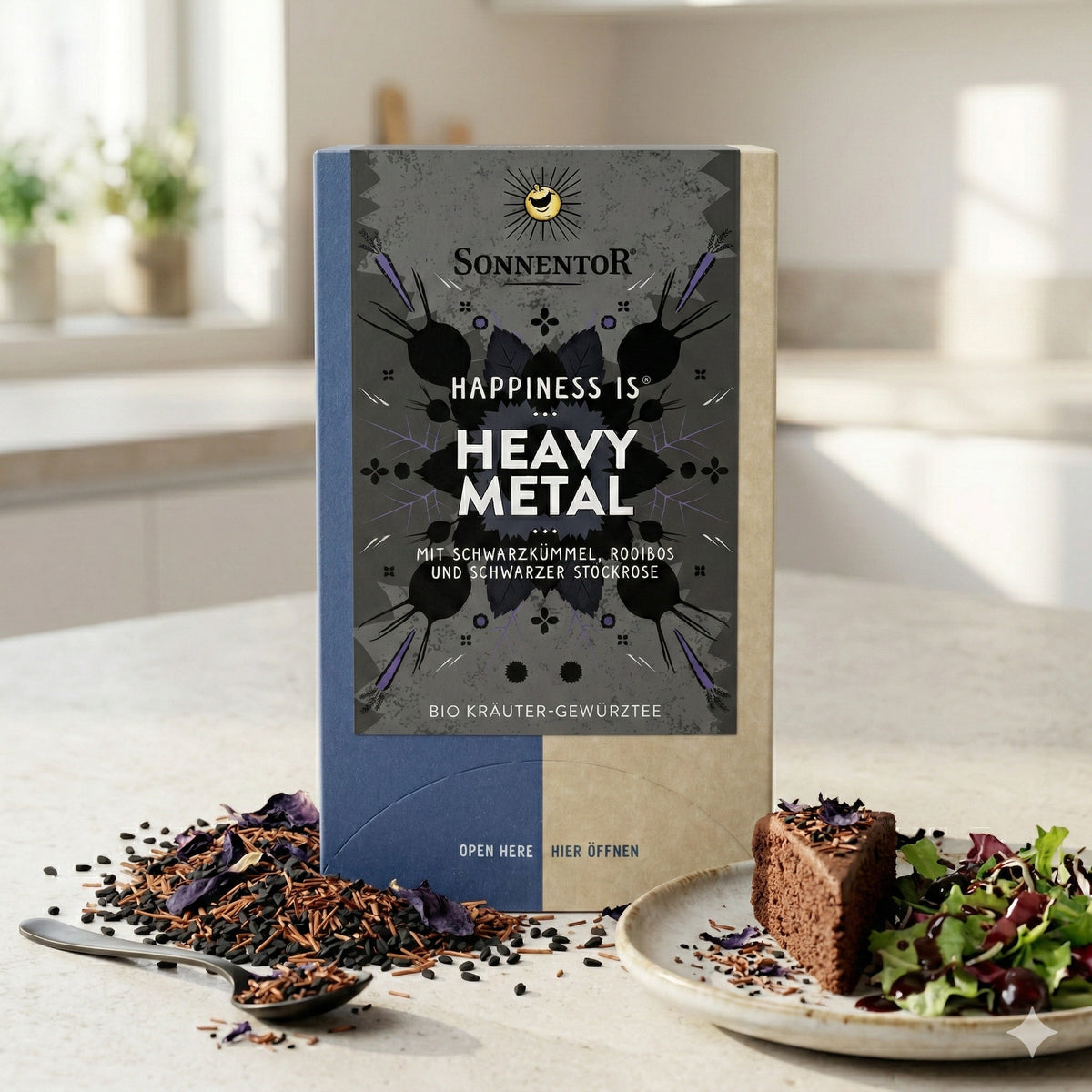Sonnentor Heavy Metal Teegetränk-Packung auf Küchenarbeitsplatte, daneben loser Tee, ein Löffel, ein Stück Kuchen und Salat auf einem Teller.