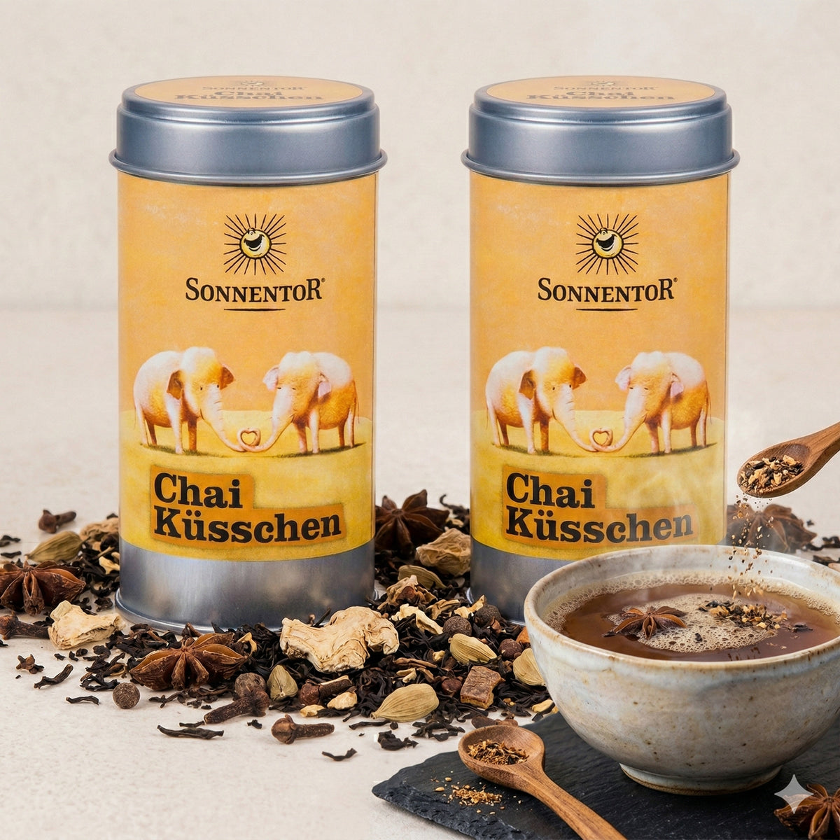 Zwei Sonnentor Chai Küsschen Gewürz-Dosen mit losen Gewürzen und einer Schale Chai Teegetränk, daneben ein Holzlöffel mit Gewürzmischungen.