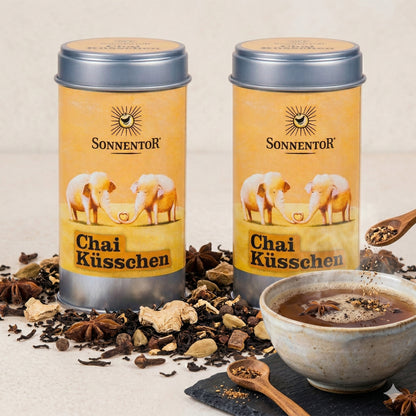 Zwei Sonnentor Chai Küsschen Gewürz-Dosen mit losen Gewürzen und einer Schale Chai Teegetränk, daneben ein Holzlöffel mit Gewürzmischungen.