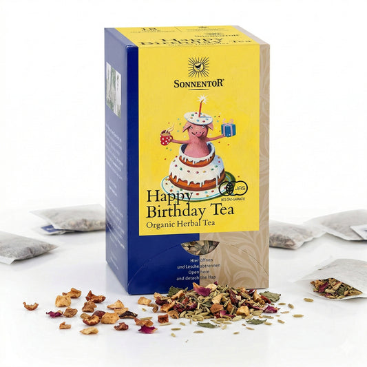 Packung Sonnentor Geburtstagstee lose mit sichtbarer Blütenmischung und Früchten vor der Verpackung, im Hintergrund Teebeutel.