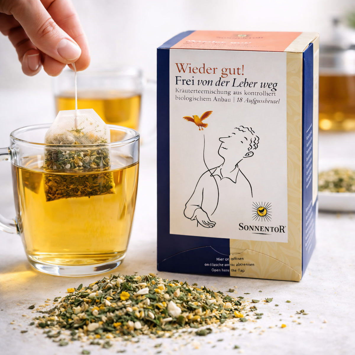 Sonnentor Tee Frei von der Leber weg neben einer Tasse mit aufgebrühtem Kräutertee, ein Teebeutel wird eingetaucht.