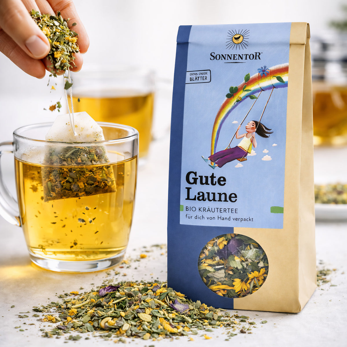 Sonnentor Gute Laune Kräutertee lose neben einer Tasse Tee, ein Teebeutel wird mit Kräutern befüllt, Kräutermischung auf dem Tisch verstreut.