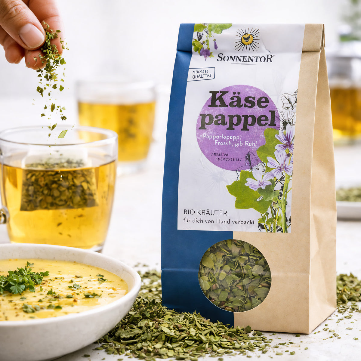 Eine Hand streut lose Käsepappel Kräuter von Sonnentor neben einer Tasse Tee und einer Schale Suppe, Packung Blaue Malve im Vordergrund sichtbar.