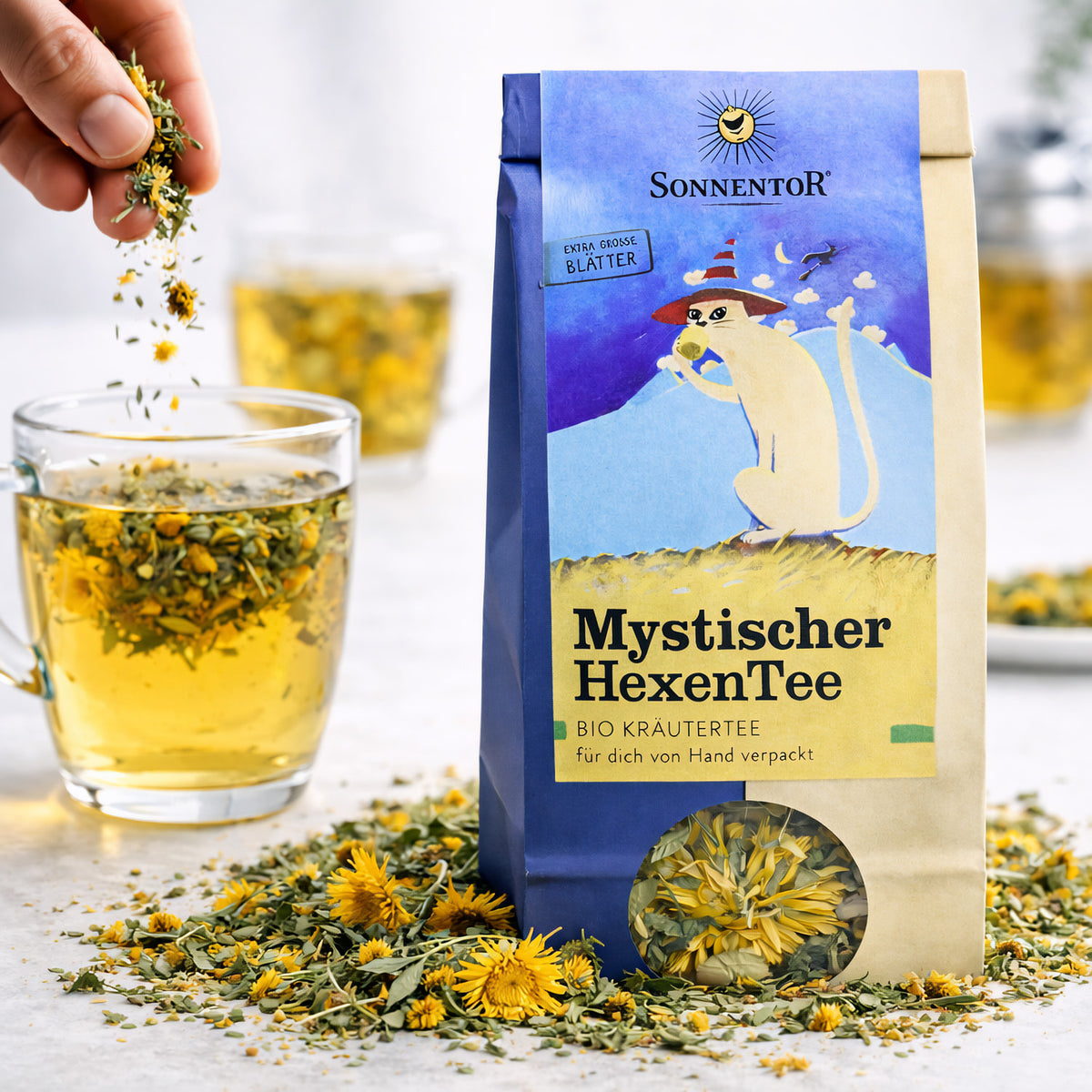Hand streut lose Kräuter des Mystischer HexenTee von Sonnentor in ein Glas mit aufgebrühtem Kräutertee, daneben die geöffnete Teepackung.