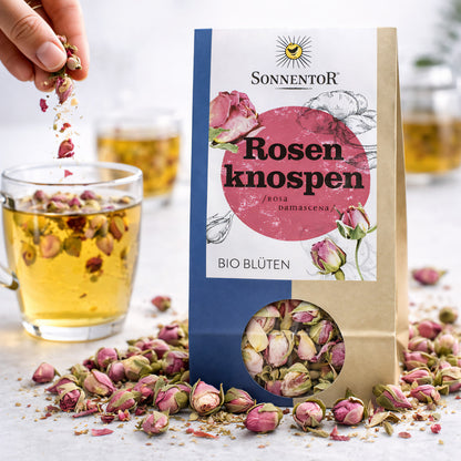 Hand streut lose Rosenknospen von Sonnentor in ein Glas mit aufgegossenem Rosentee, Packung und Kräuter im Vordergrund.