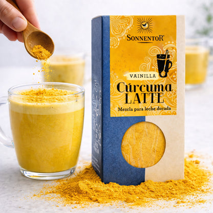 Sonnentor Kurkuma Latte Vanilla Gewürzmischung neben einem Glas mit gelbem Latte, Hand streut mit Holzlöffel Kurkuma-Pulver auf das Getränk.