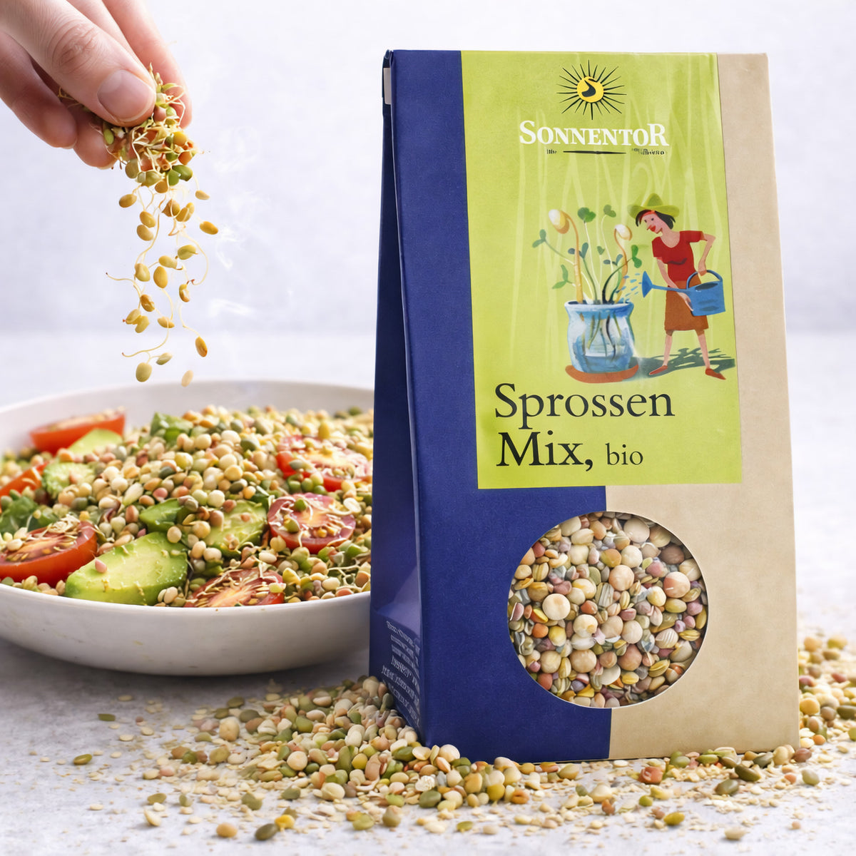 Sonnentor Sprossen-Mix 120g neben einer Schale Salat, Hand streut Keimsprossen und Saaten über das Gericht, Verpackung im Vordergrund.