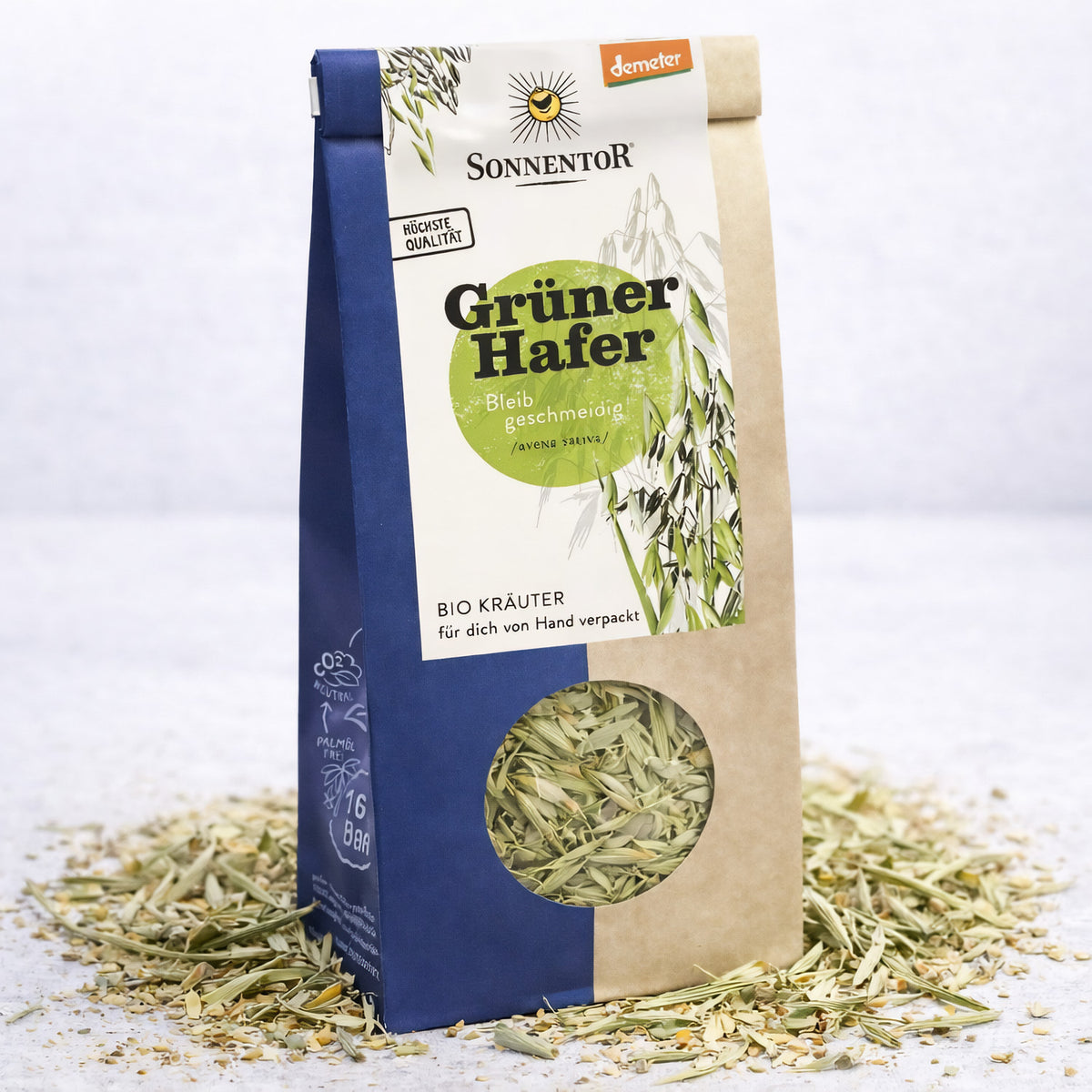 Packung Sonnentor Grüner Hafer Kräuter lose stehend, umgeben von verstreutem Hafer auf hellem Untergrund.