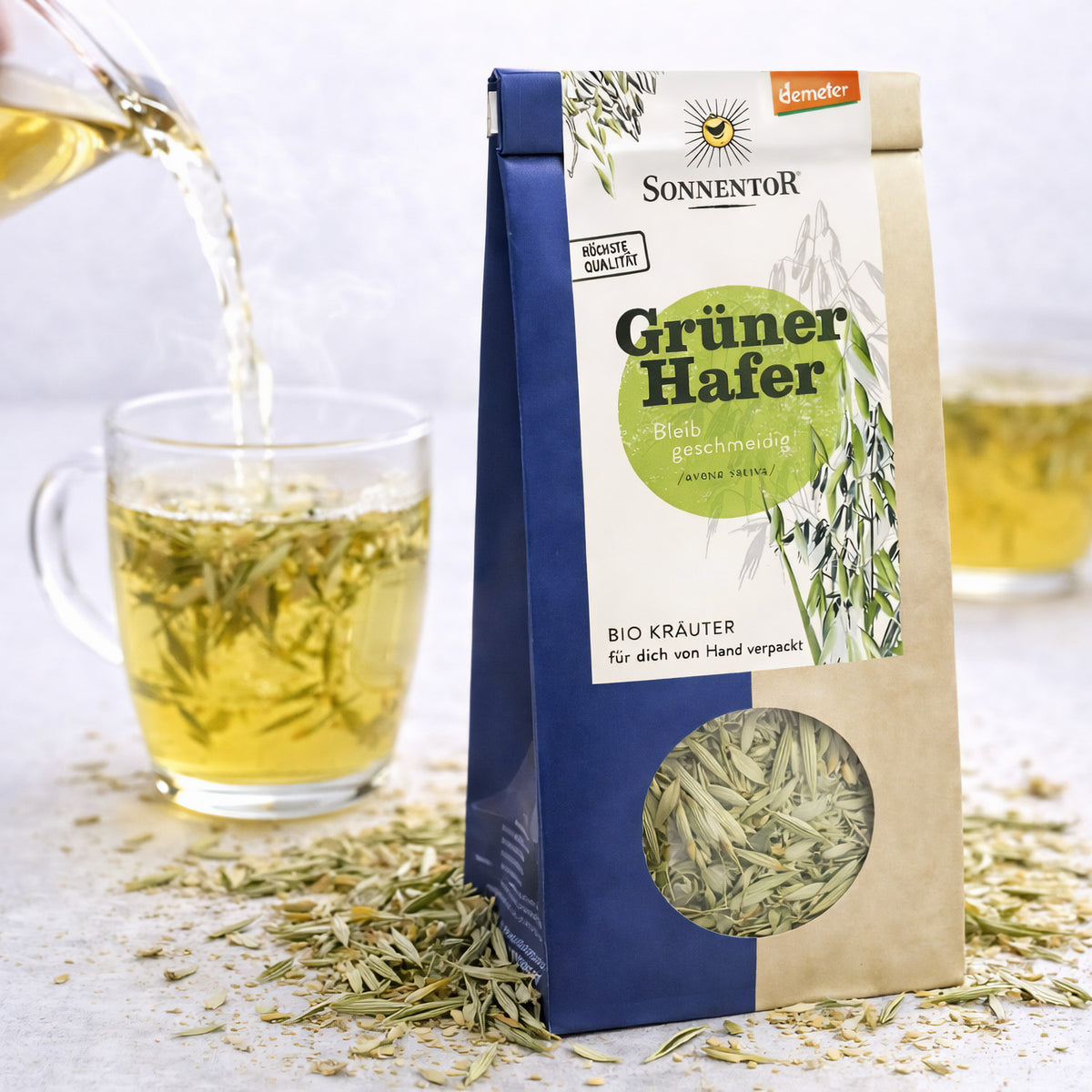 Sonnentor Grüner Hafer Tee neben einer Tasse, in die gerade Kräutertee aus einer Kanne gegossen wird.