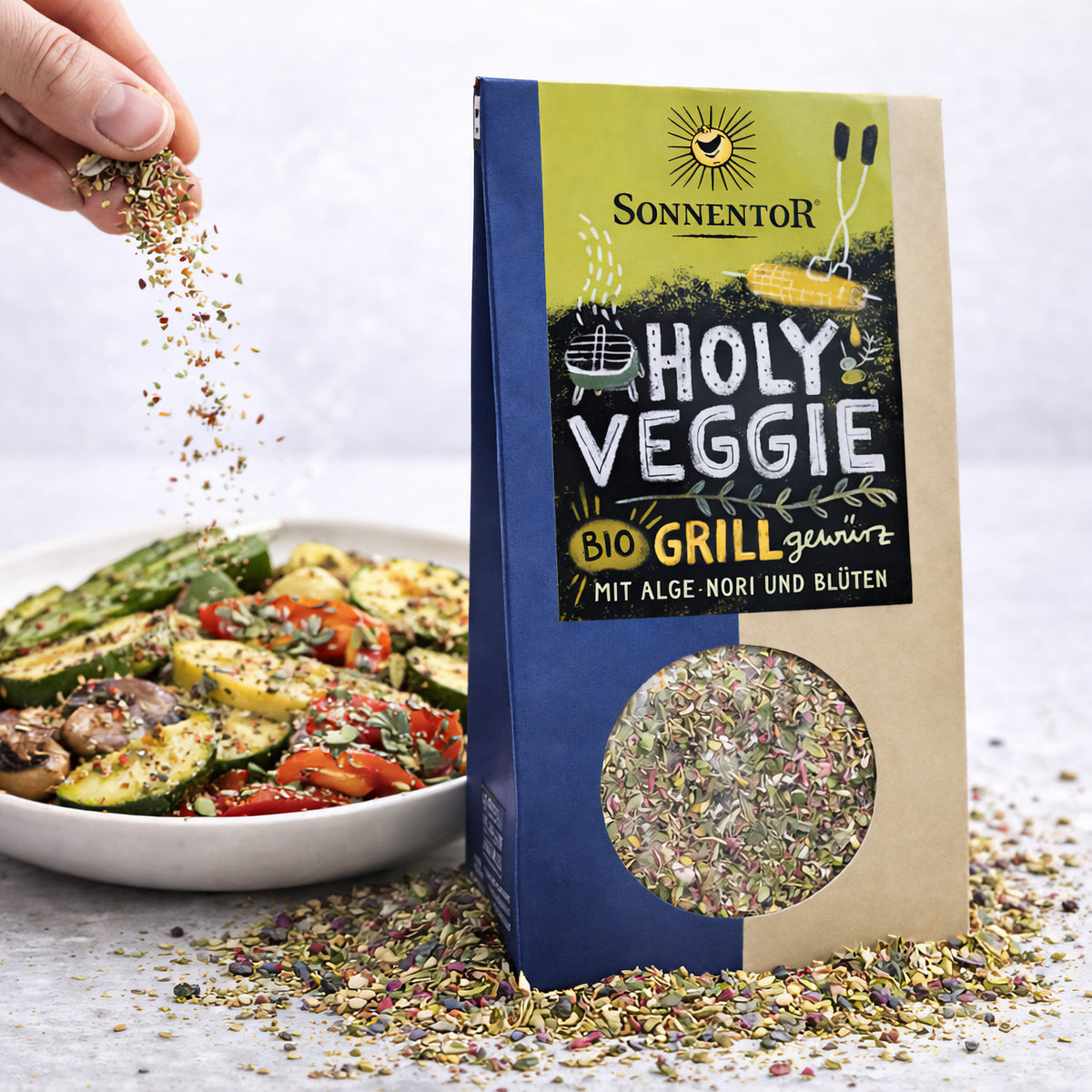 Sonnentor Holy Veggie Grillgewürz neben einem Teller Grillgemüse, eine Hand streut das bunte Gewürz über das Gemüse.