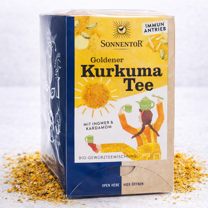 Packung Sonnentor Goldener Kurkuma Tee Doppelkammerbeutel mit gelbem Design und illustriertem Motiv, umgeben von verstreutem Kurkuma.