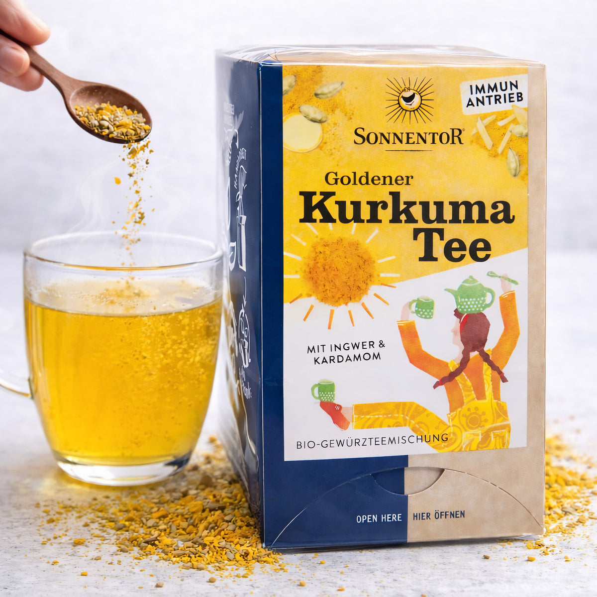 Sonnentor Goldener Kurkuma Tee neben einer Tasse mit aufgebrühtem Tee, ein Löffel Kurkuma-Gewürzmischung wird in die Tasse gegeben.