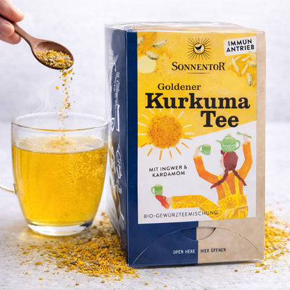 Sonnentor Goldener Kurkuma Tee neben einer Tasse mit aufgebrühtem Tee, ein Löffel Kurkuma-Gewürzmischung wird in die Tasse gegeben.