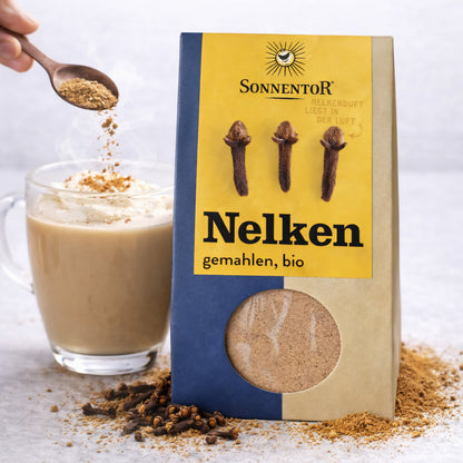Sonnentor Nelken gemahlen neben einer Tasse mit Milchschaum, darüber wird das Gewürz mit einem Löffel gestreut.