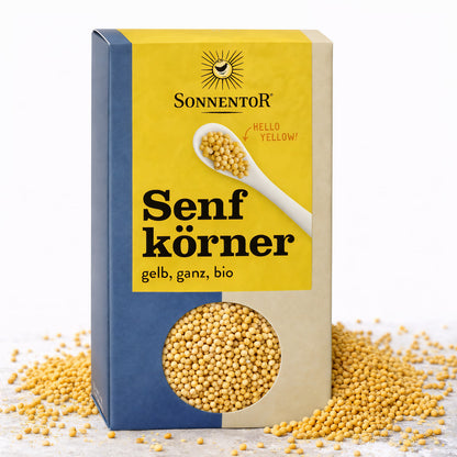 Packung Sonnentor Senfkörner gelb ganz mit Sichtfenster, umgeben von verstreuten Senfsaat-Körnern auf hellem Untergrund.